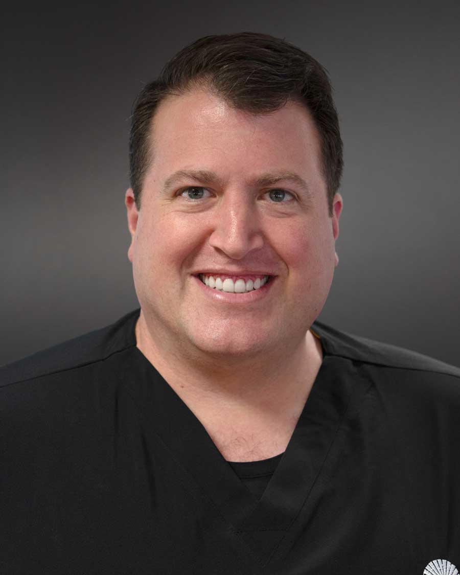 The image features a man wearing a white shirt with a black tie, smiling at the camera, posing for a portrait-style photograph against a dark background.