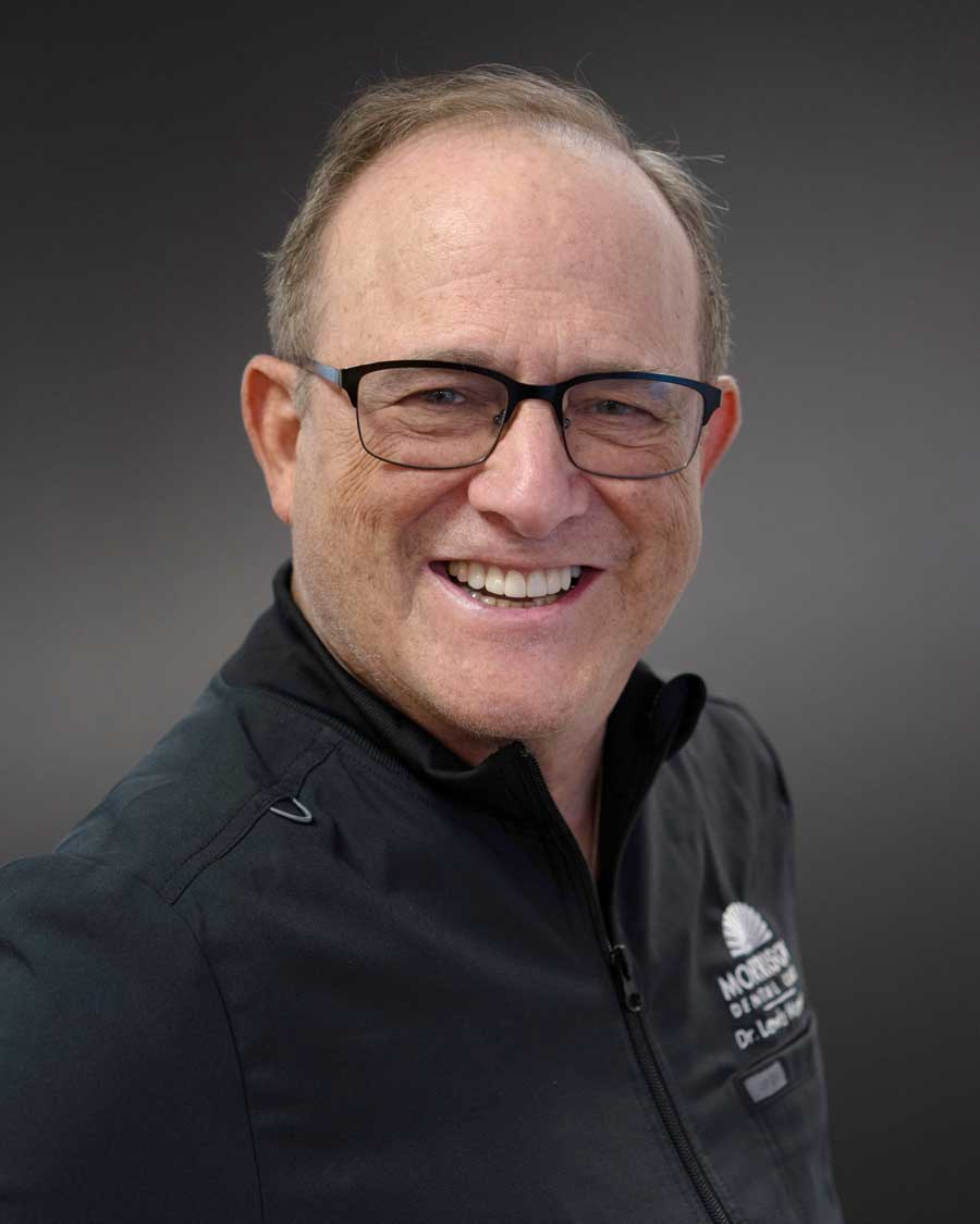 The image features an older man with glasses smiling at the camera, wearing a dark jacket and light-colored shirt, against a dark background.