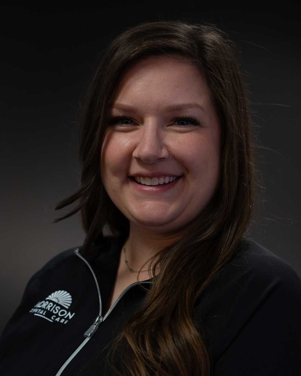 The image shows a smiling woman with long hair, wearing a black jacket with a logo on the left side, posing against a dark background.