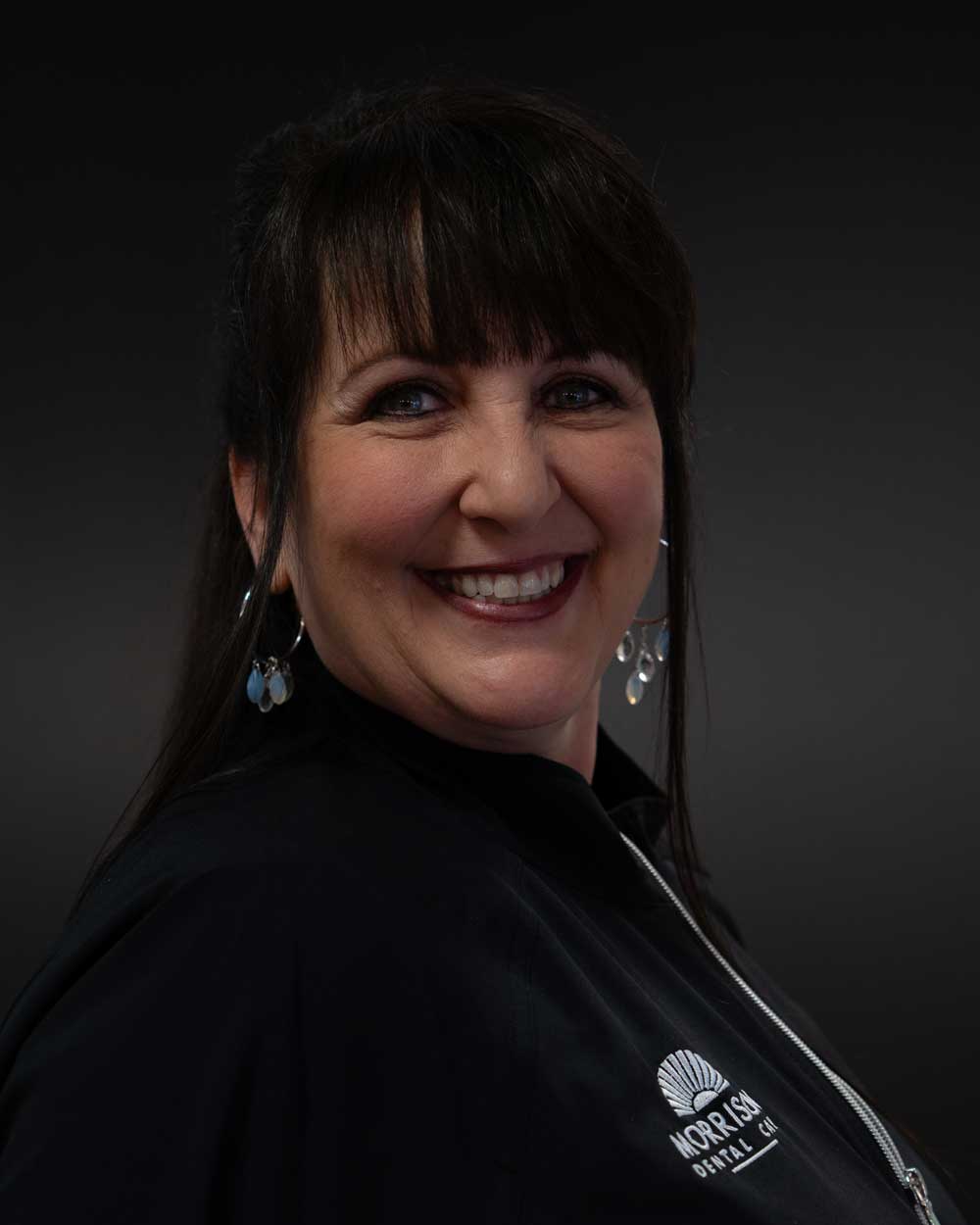 The image shows a woman with short hair wearing a black top and earrings, standing against a dark background.