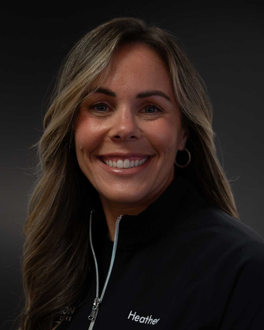 The image shows a woman with a broad smile, wearing a black jacket with white detailing, a name tag, and a headset microphone, posing against a dark background.