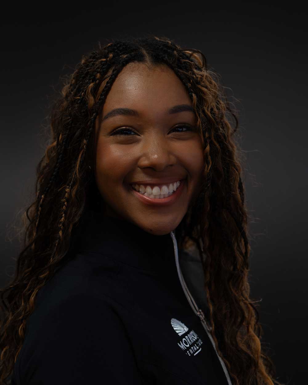The image shows a smiling woman with braided hair, wearing a black top with a logo on the left side, against a dark background.