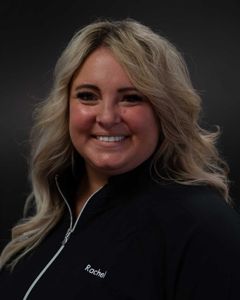The image shows a woman with blonde hair smiling at the camera, wearing a black jacket with a white zipper, and a name tag on her right side.