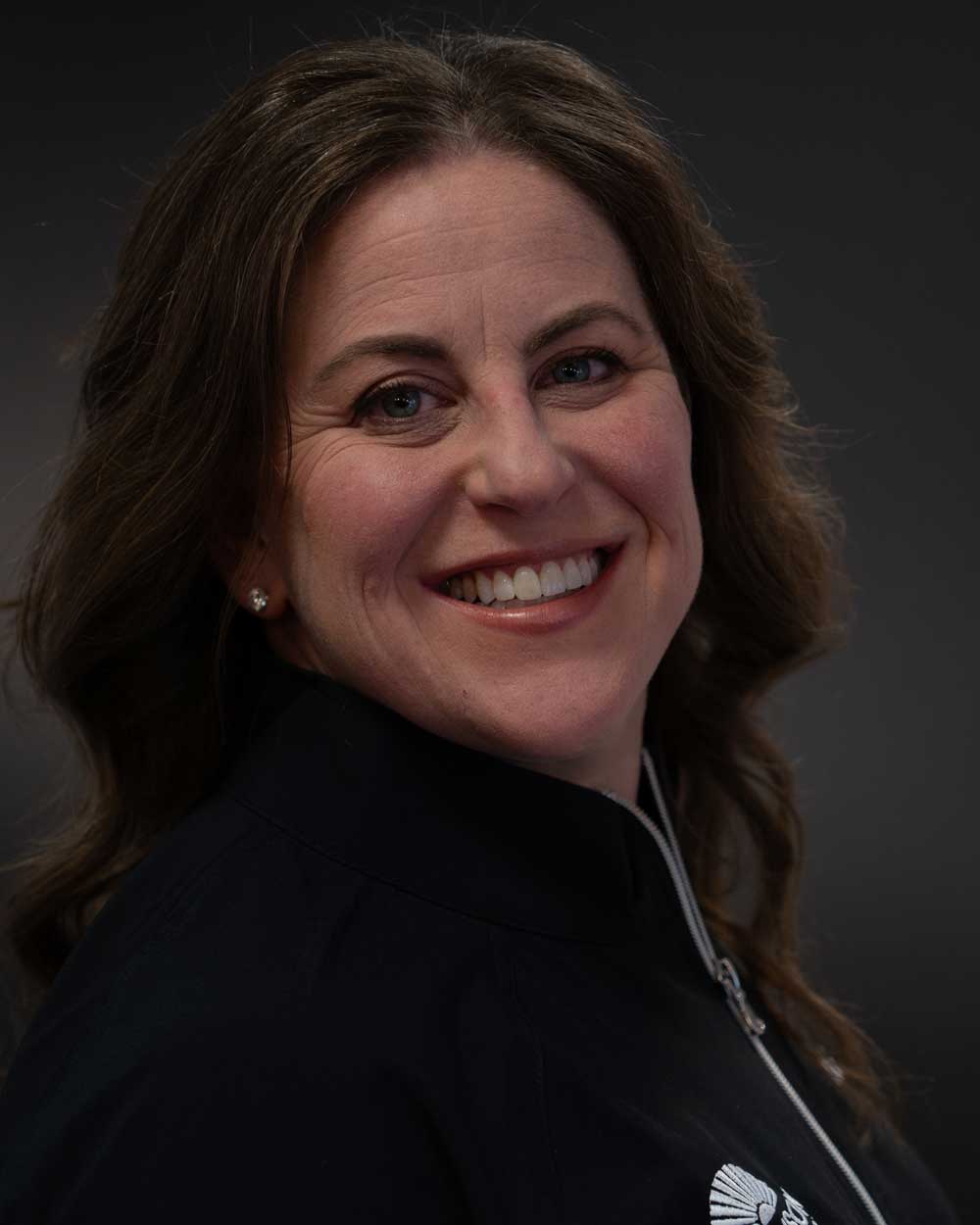 The image shows a woman with a friendly smile posing for a portrait, wearing a black jacket with a logo on the left chest area and a white lanyard around her neck.