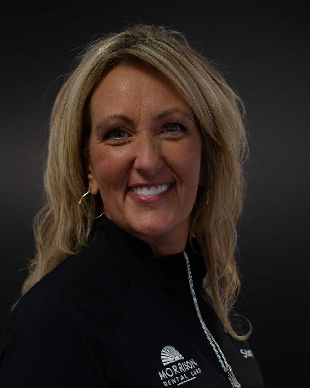 The image shows a woman with blond hair, wearing a black jacket with a logo on the left side, smiling at the camera against a dark background.